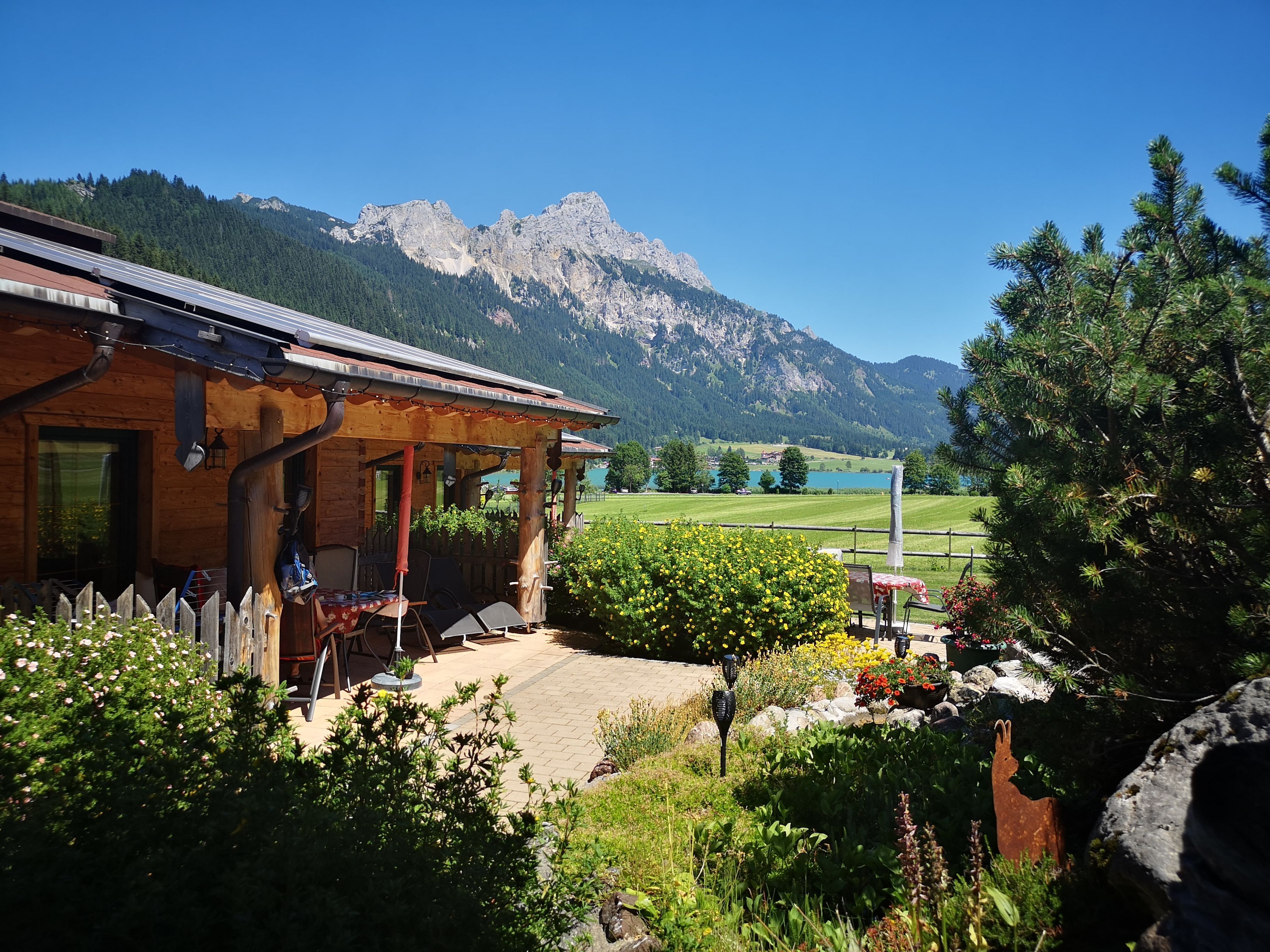 Een houten chalet met een overdekt terras, uitzicht op een bergketen en een verzorgde tuin met verschillende planten en bloemen.