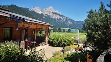 Een houten chalet met een overdekt terras, uitzicht op een bergketen en een verzorgde tuin met verschillende planten en bloemen.