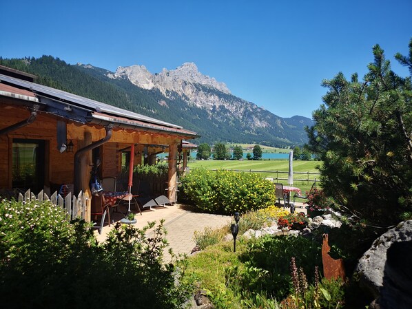 A wooden cabin with a covered patio, a view of a mountain range, and a well-maintained garden with various plants and flowers.