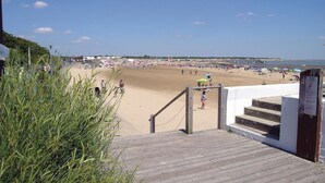 Una playa con una escalera de madera que baja a la arena, un cielo despejado y un muelle a lo lejos.