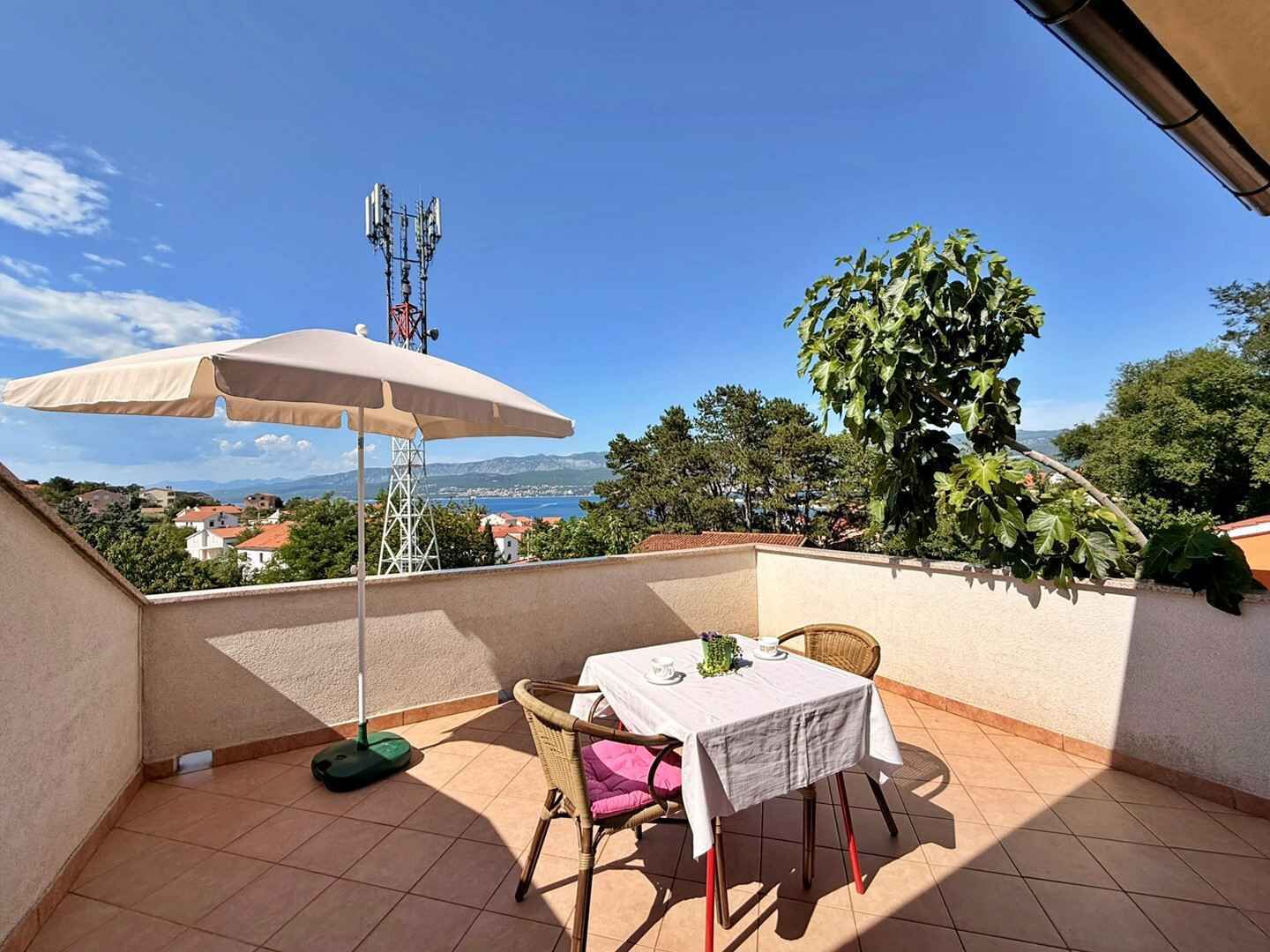 Un balcón con mesa, sillas y sombrilla, con vistas panorámicas a un pueblo y montañas.