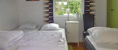 A bedroom with two beds, a window with a view of greenery, a radiator, and a small cabinet.