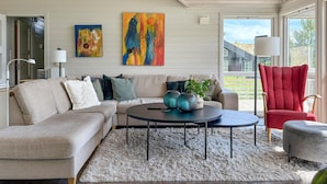 Un salon moderne avec un canapé d’angle beige, une table basse ronde et un fauteuil rouge. Deux tableaux abstraits ornent le mur, et une plante en pot est posée sur la table basse.