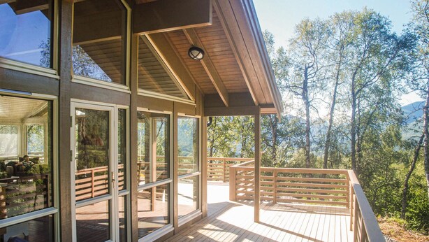 Une terrasse en bois avec des portes vitrées donnant sur une pièce, le tout entouré d’arbres et avec une chaîne de montagnes en arrière-plan.