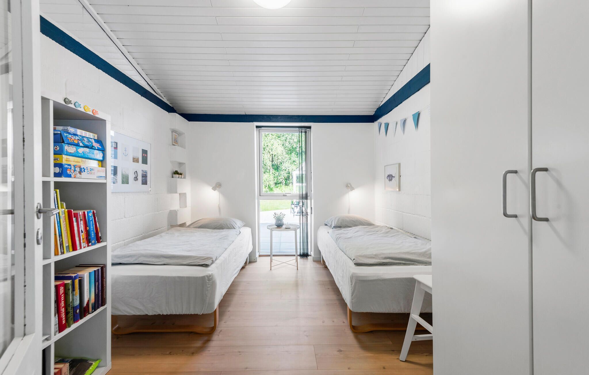 A bedroom with two single beds, a bookshelf with books, a small table with a lamp, and a window with a view of greenery.