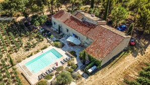 Un bâtiment en pierre avec un toit en tuiles, une piscine avec des chaises longues et une zone boisée.