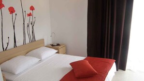 Una cama bien tendida con ropa blanca y roja, una mesita de noche con lámpara y una pared con diseño floral.