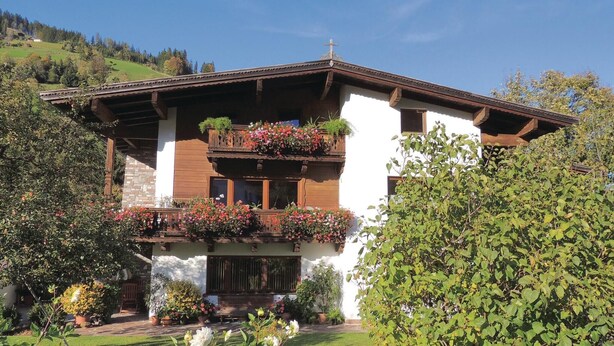 A traditional chalet with a balcony adorned with flowers, a stone foundation, and a cross on the roof.
