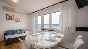 Un coin repas avec une table blanche dressée pour quatre personnes, des chaises blanches, un canapé avec des coussins bleus et une fenêtre donnant sur la mer.