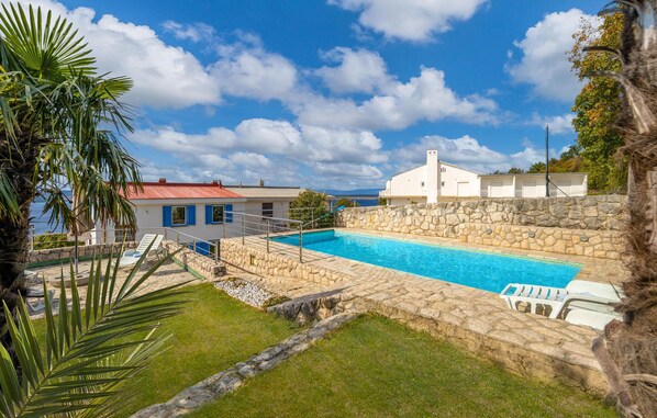 A swimming pool with stone walls, a grassy area, and a building with blue shutters.