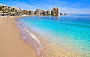 Una playa con agua turquesa cristalina, una orilla arenosa y una fila de edificios de varios pisos al fondo.