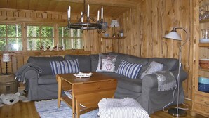 A cozy cabin interior with a wooden ceiling, a gray sectional sofa with striped pillows, a wooden coffee table, a floor lamp, and a window with a view of trees.