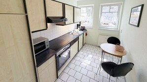 Una cocina compacta con horno empotrado, microondas y cafetera sobre la encimera. Hay una mesita con una silla y una cesta.