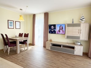Una sala de estar moderna con comedor, un televisor de pantalla plana en la pared, un mueble de madera con un radio y una ventana con cortinas.