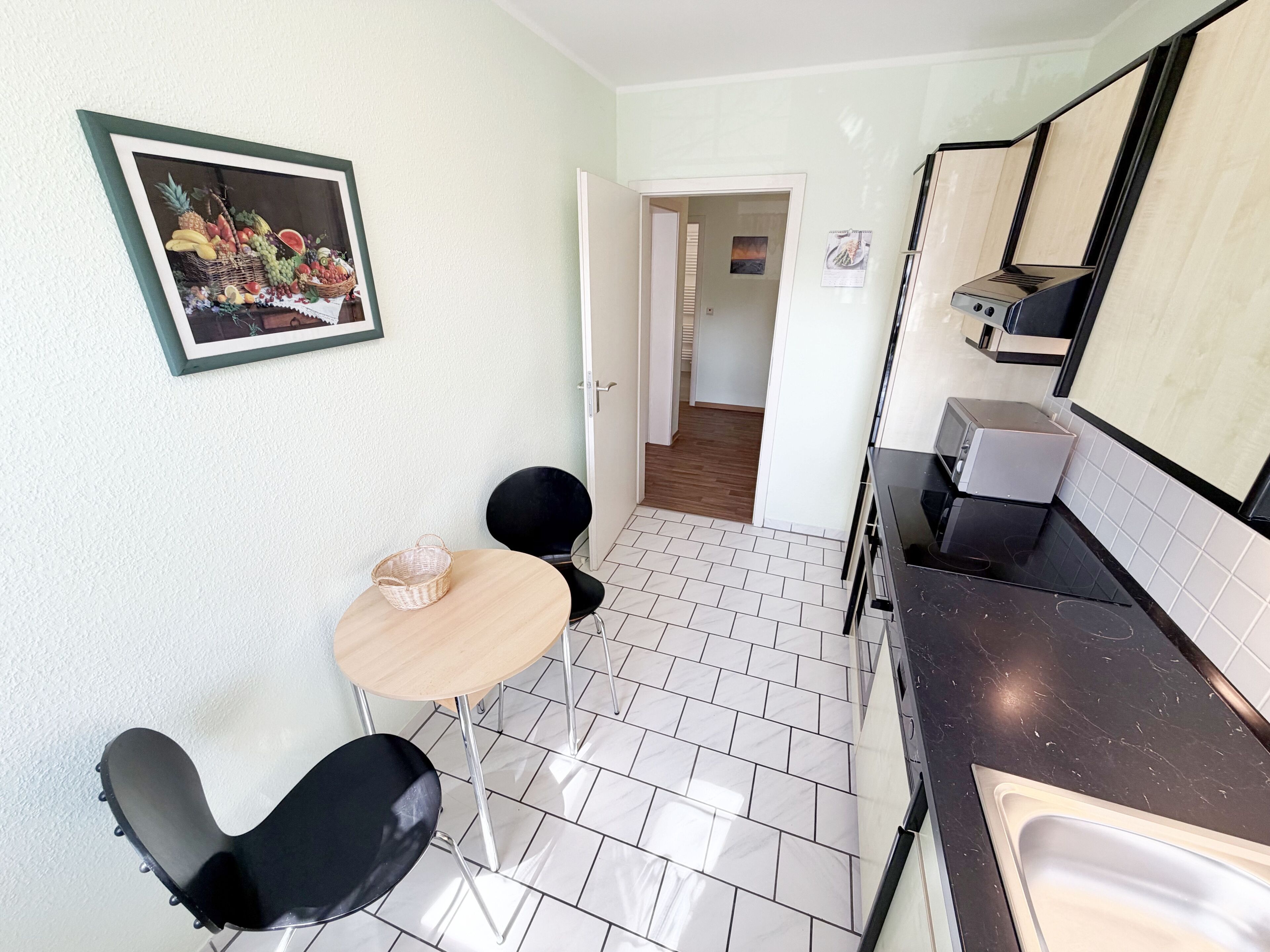 Una pequeña kitchenette con un lavamanos, estufa y microondas. Hay una mesa pequeña con dos sillas y una cesta encima. Un cuadro enmarcado de frutas está en la pared.