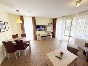A modern living room with a dining area, a flat-screen TV, and a large window with curtains.