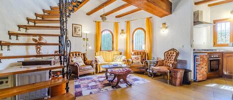 A spacious living room with a wooden staircase, two large windows with curtains, a fireplace, and a kitchen area.