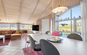 Ein gemütliches Zimmer mit Holzbalkendecke, einem großen Fenster mit Blick ins Grüne, einem Esstisch mit Stühlen und einem Flachbildfernseher.