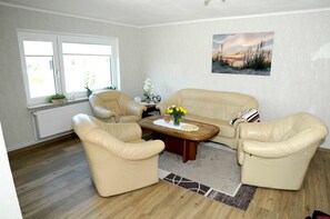 Ein Wohnzimmer mit einem beigen Sofa, zwei Sesseln, einem Holztisch und einem Fenster mit Blick ins Freie.