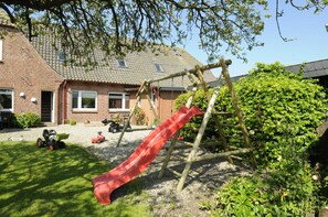A playground with a slide, surrounded by a brick house, a lawn, and a hedge.