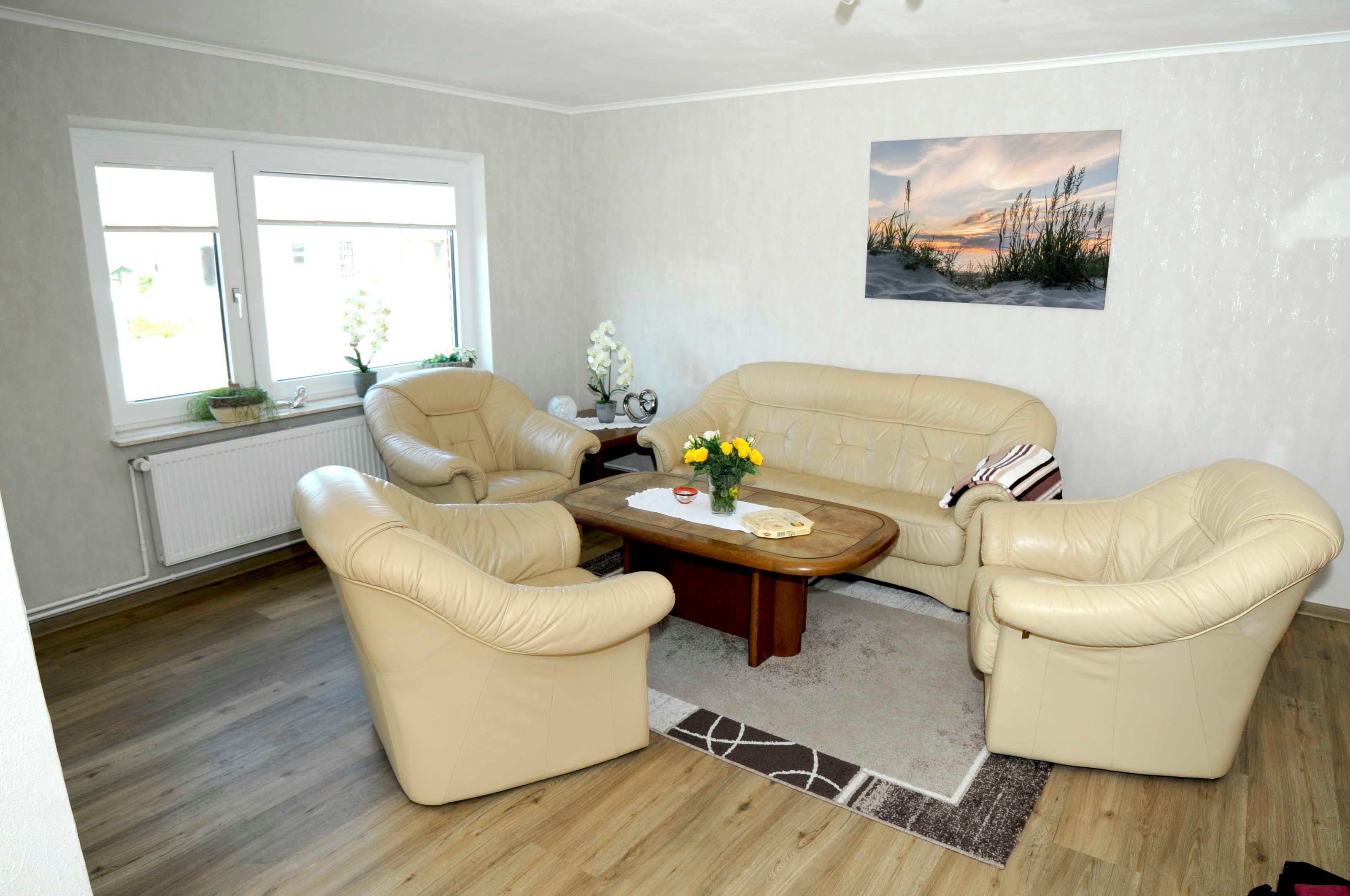 Ein Wohnzimmer mit einem beigen Sofa, zwei Sesseln, einem Holztisch und einem Fenster mit Blick ins Freie.