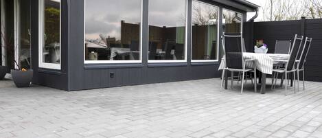 A modern house with large windows, a patio with a table and chairs, and a flower planter.