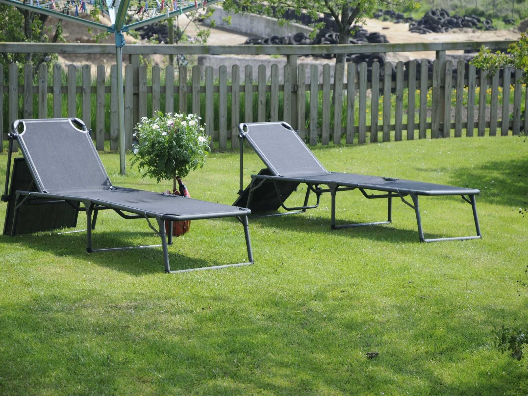 Two outdoor lounge chairs with metal frames and fabric seats, positioned on a grassy lawn with a wooden fence in the background.