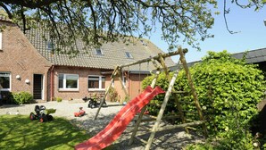 A playground with a slide, surrounded by a brick house, a lawn, and a hedge. - Vacation apartment Meer, Germany (Hasselberg)