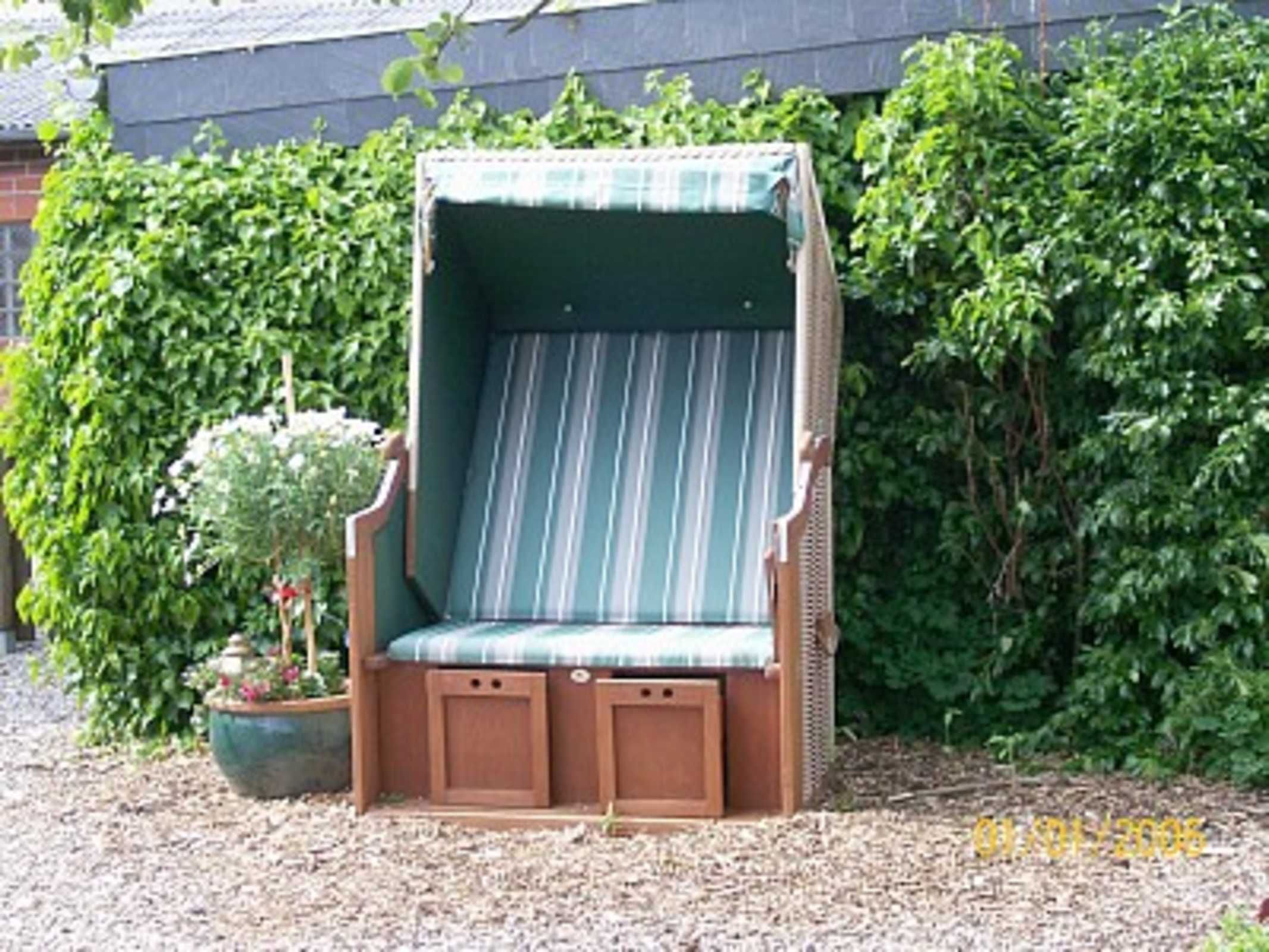 Ein Holzstuhl im Freien mit gestreifter Sitzkissen, ein kleiner Blumentopf und ein dichter, grüner Heckenstreifen im Hintergrund.