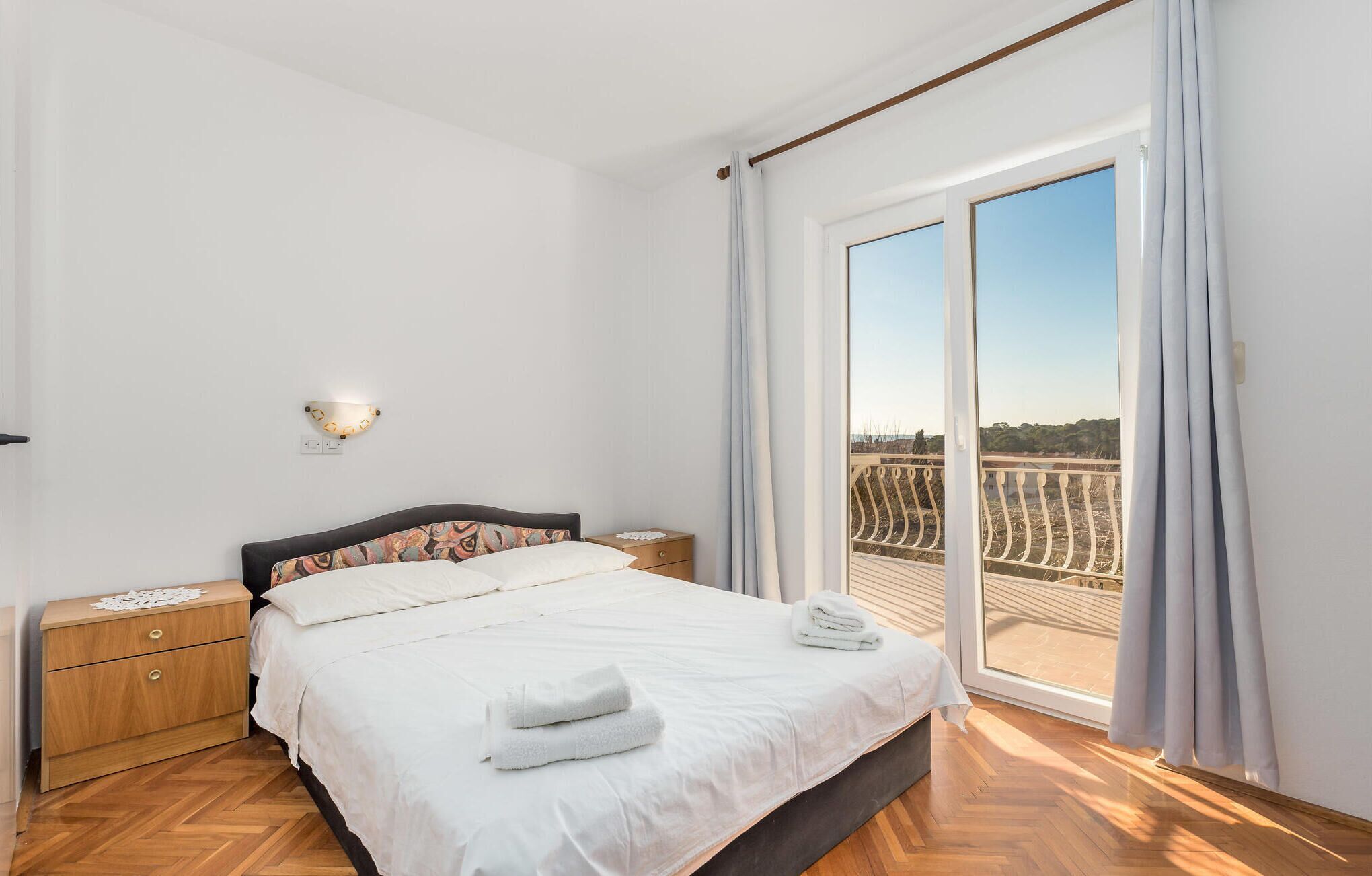 A bedroom with a bed, a nightstand, a wall-mounted light, and a large window with a view of a balcony.