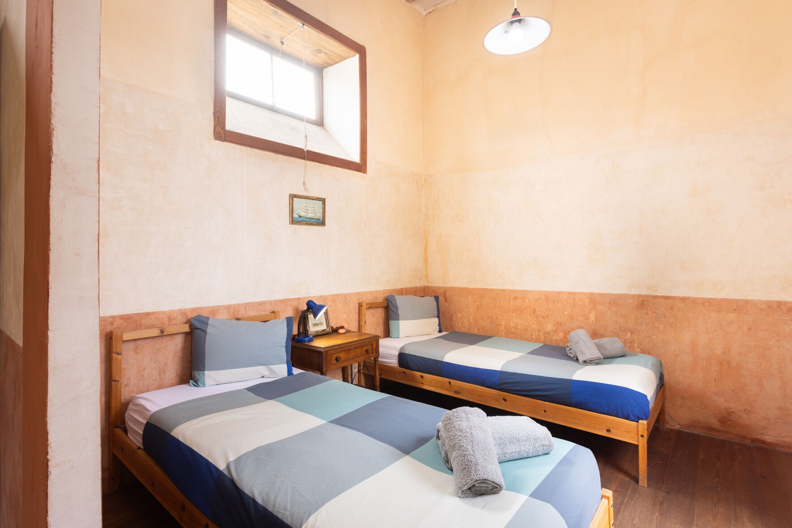 Ein Zimmer mit zwei Holzbetten, einem kleinen Tisch, einem Nachttisch, einem Fenster und einer Pendelleuchte.