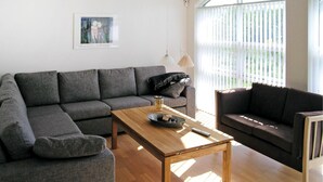 Ein modernes Wohnzimmer mit einem grauen Ecksofa, einem dunklen Sessel und einem Holztisch. An der Wand hängt ein Gemälde und ein Fenster ist mit Jalousien versehen.