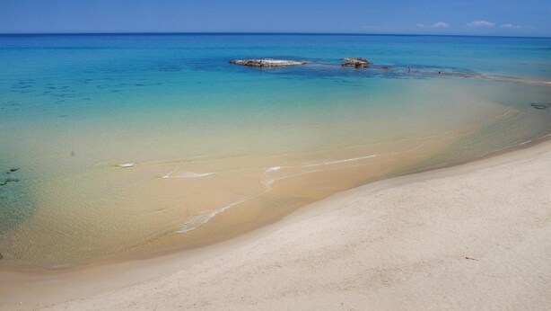 Een strand met helder turquoise water, een zandstrand en een rotsachtige landtong in de verte.