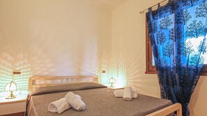 Een net opgemaakt bed met twee opgerolde handdoeken, een bedlampje en een raam met een blauw gordijn.