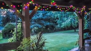 Un espacio al aire libre cubierto con estructura de madera, luces colgantes y vistas a un paisaje con césped y árboles.