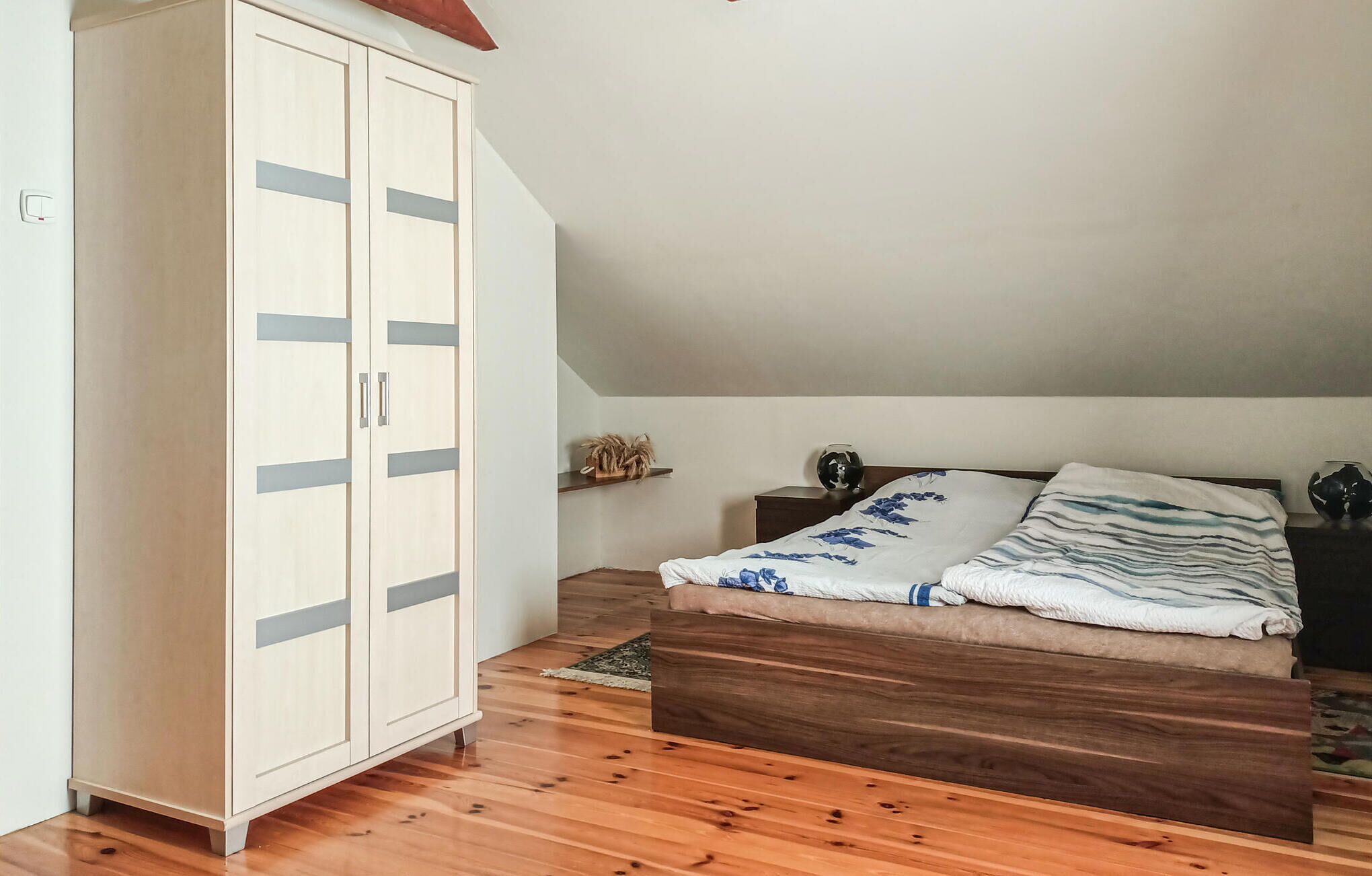 Une chambre avec un plancher en bois, un lit recouvert d’une couvre-lit à motifs bleus et blancs, une armoire en bois et une petite étagère ornée d’objets décoratifs.