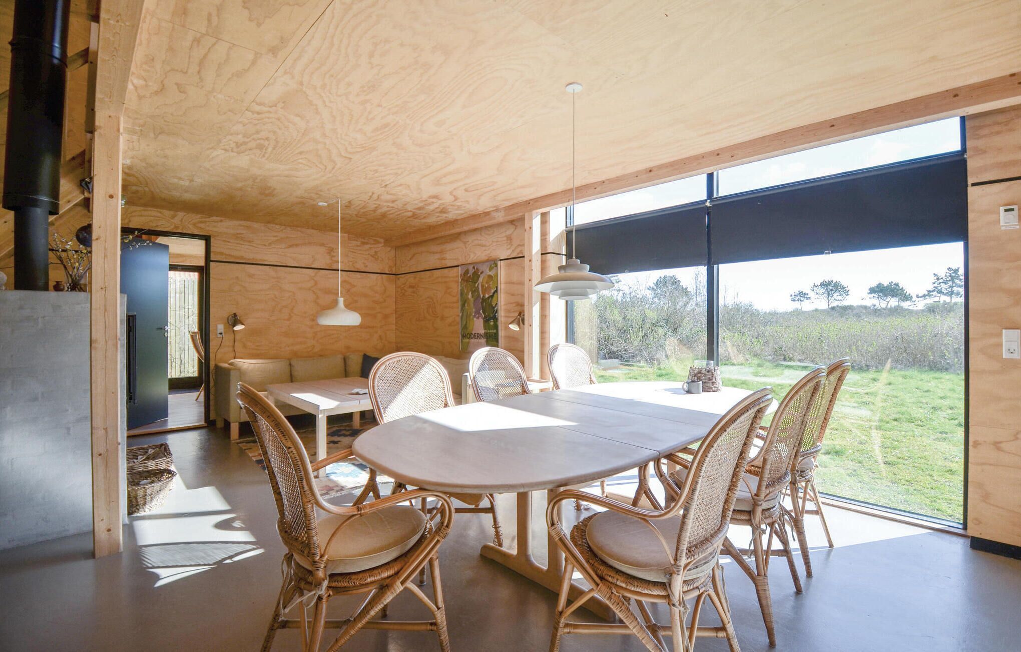 A modern wooden interior with a dining area featuring a round table and wicker chairs, large windows offering a view of the outdoors, and a minimalist design.