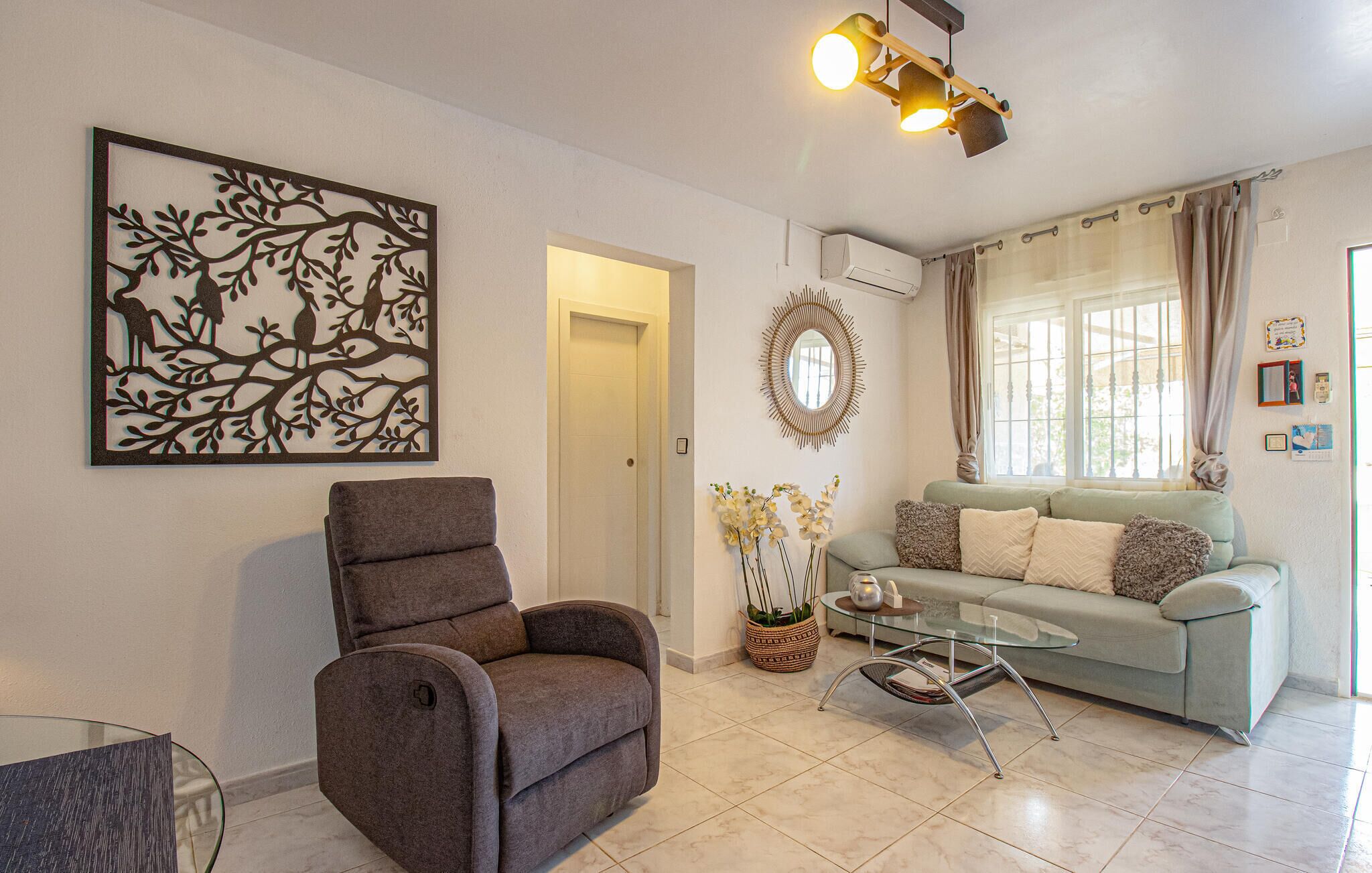 Ein Wohnzimmer mit grauem Sofa, braunem Sessel, Glastisch und einem wandmontierten Spiegel.