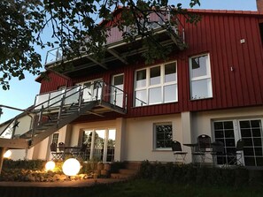 A two-story building with a red exterior, a balcony with metal railings, and a patio area with outdoor seating and lighting. - Apartment 5 - Usedom Landhaus Morgenitz (Mellenthin OT Morgenitz)