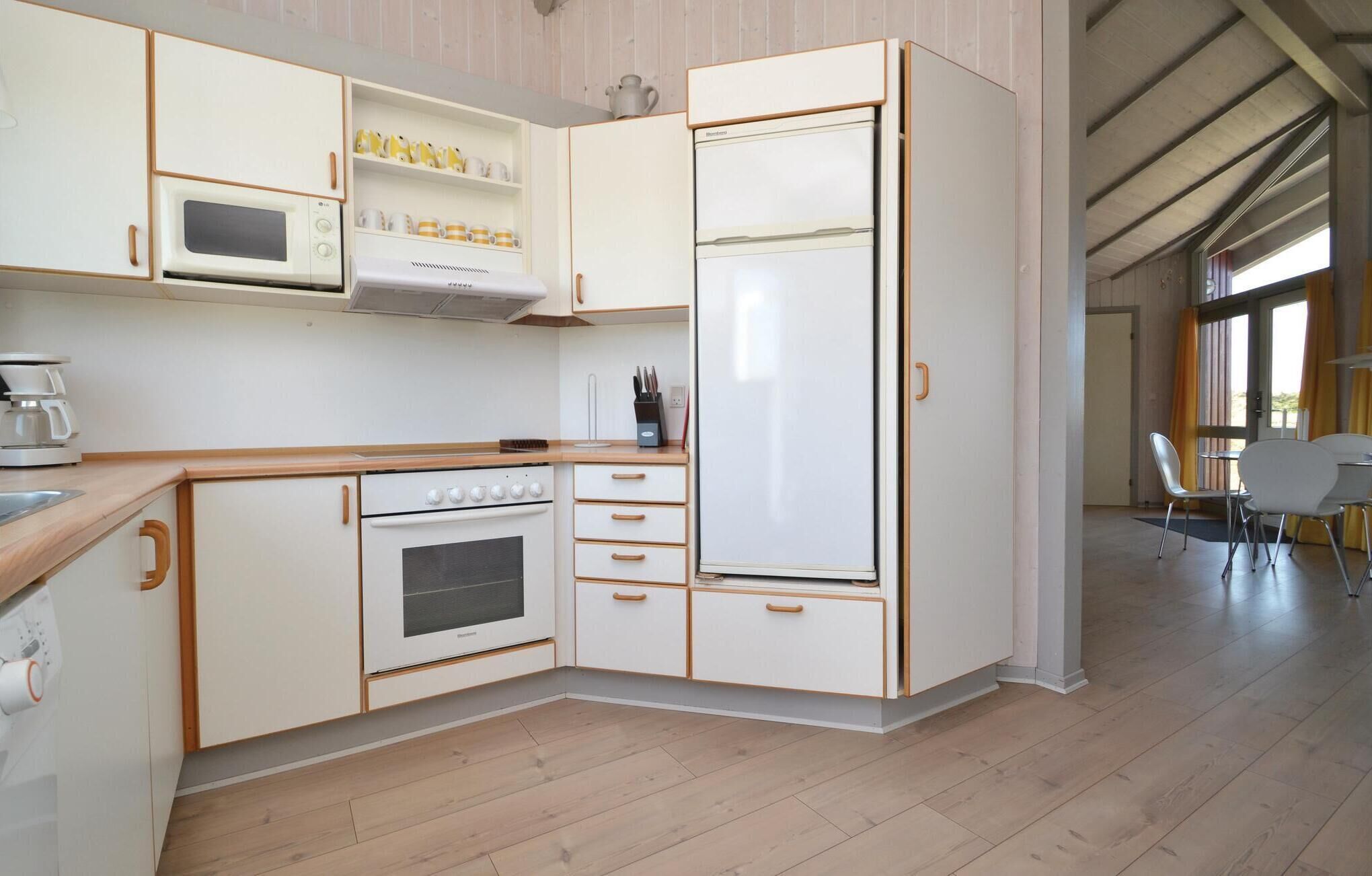 Cocina compacta con muebles blancos, horno integrado, microondas y refrigerador. Cuenta con encimera de madera y un área de comedor con sillas a la vista al fondo.