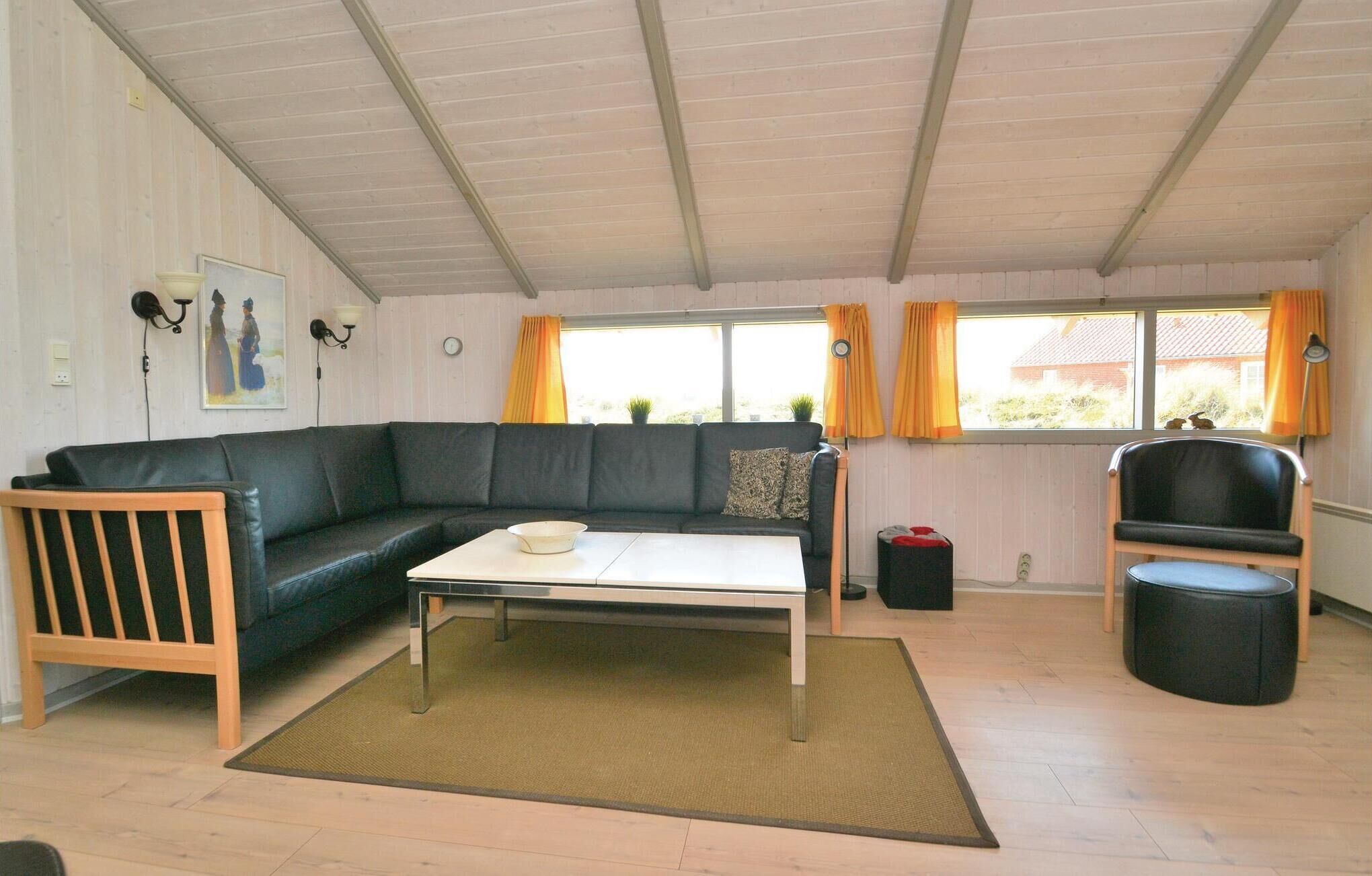 Un salon comprenant un canapé d’angle, une table basse, une chaise en bois, un pouf noir et une fenêtre avec des rideaux.