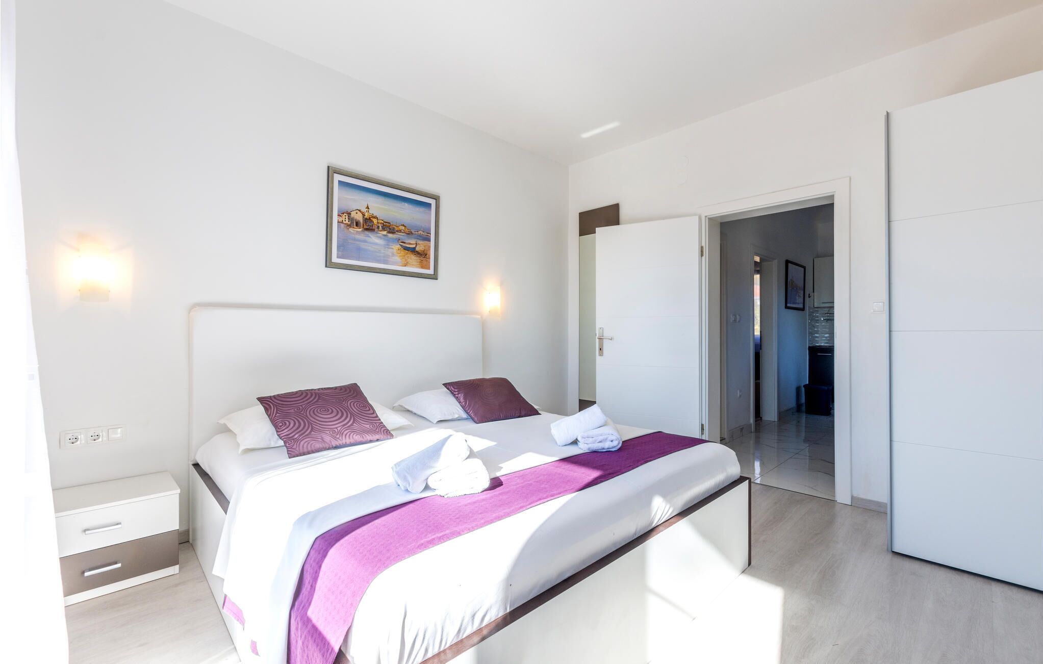 Un dormitorio moderno con cama, mesitas de noche, un cuadro y vistas a un pasillo.