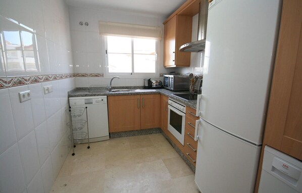 A compact kitchen with white tiles, wooden cabinets, a built-in oven, a microwave, and a refrigerator.