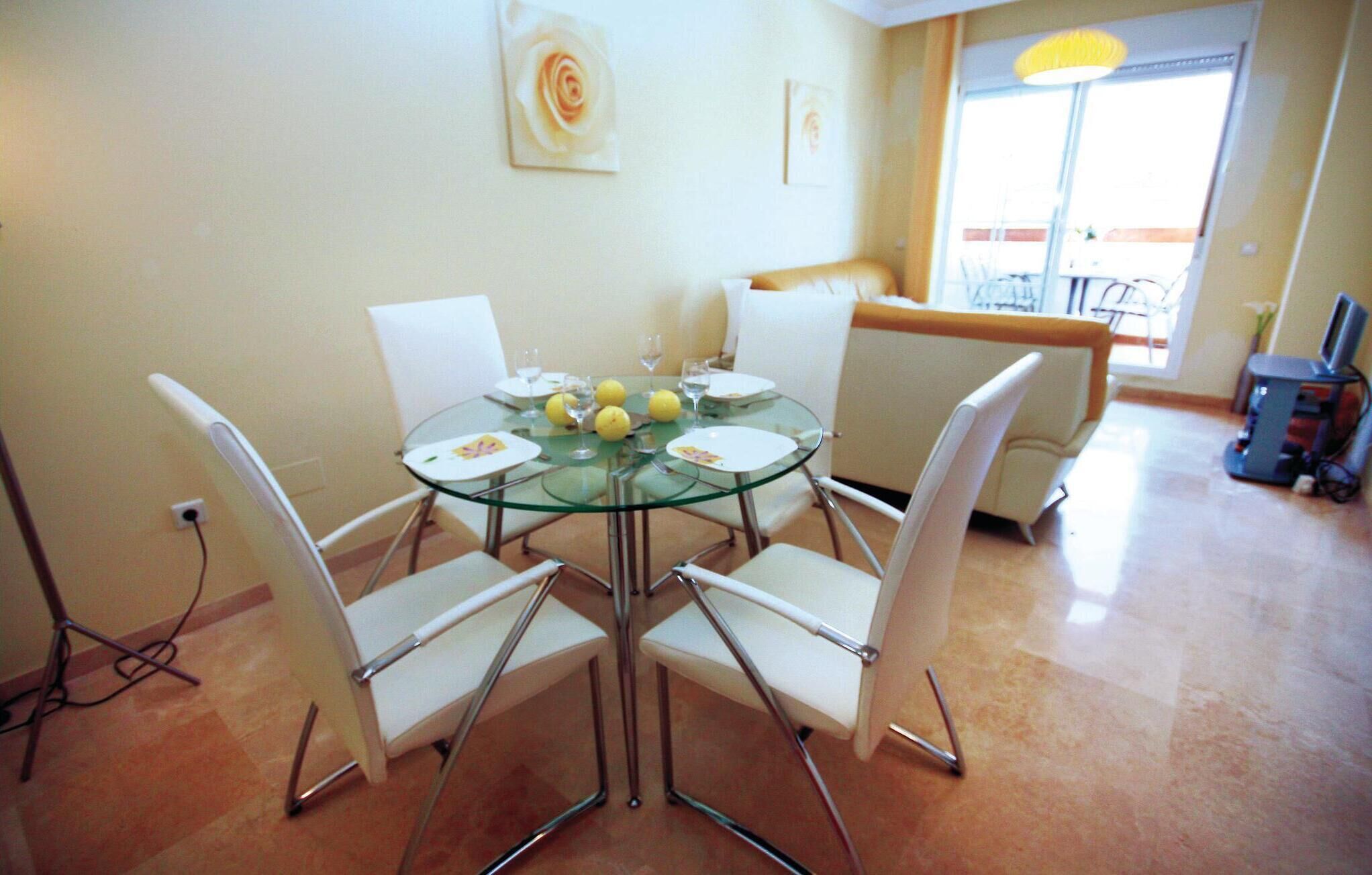 A dining area with a glass table surrounded by white chairs, a sofa, and a television in the background.