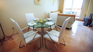 Un coin repas avec une table en verre entourée de chaises blanches, un canapé et un téléviseur en arrière-plan.