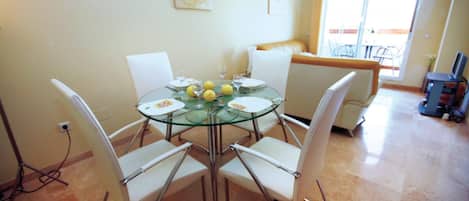 A dining area with a glass table surrounded by white chairs, a sofa, and a television in the background.