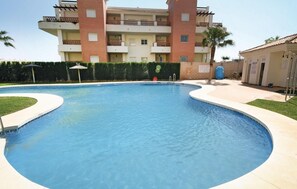 A swimming pool with a curved edge, surrounded by a low hedge and a building with balconies in the background.