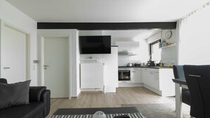 Una sala moderna con un sofá negro, una mesa de centro con un control remoto, y una zona de cocina con armarios blancos y un televisor de pantalla plana.