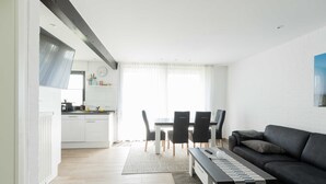 Una sala de estar moderna con un sofá negro, una mesa de comedor con sillas, una zona de cocina con armarios blancos y un ventanal con cortinas translúcidas.