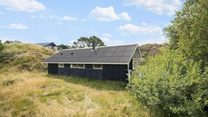 A small, dark-colored cabin with a sloped roof, surrounded by grass and shrubs, with a clear sky above. - 2 bedroom nice home in Fanø (Fanø)